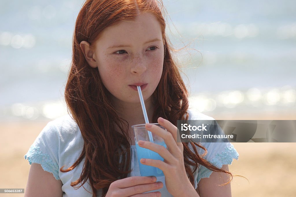 Girl drinking slushie drink with straw on beach, frozen ice-drink Photo showing a girl with long red hair and a summer T-shirt, drinking a slushie ice drink with a straw.  The girl is pictured in the full sunshine, standing on a beach while she drinks her blue, raspberry flavoured ice / frozen drink, with the lapping waves of the sea sparkling in the background. Slushie - Drink Stock Photo