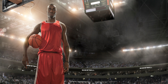 Professional basketball player standing holding basketball on an indoor floodlit basketball court full of spectators.