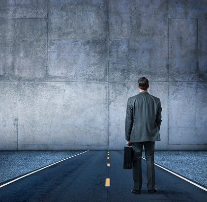 A businessman, with his back to the camera while holding a briefcase, stands in the middle of a road that runs directly into a large concrete wall.