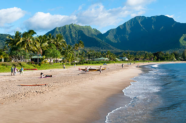 Paddle boarder and red outrigger canoe off the coast of Puu Poa Beach with mountains in the background on a sunny day with blue sky and clouds, Princeville, Kauai, Hawaii