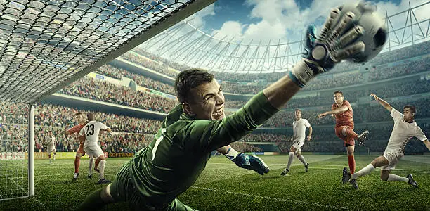 View from inside of the soccer gates - a male soccer goalie jumping in motion for a ball while defending his gates on wide angle panoramic image of a outdoor soccer stadium or arena full of spectators under a sunny sky. The image has depth of field with the focus on the foreground part of the pitch. With intentional lensflares. Player is wearing unbranded soccer uniform.