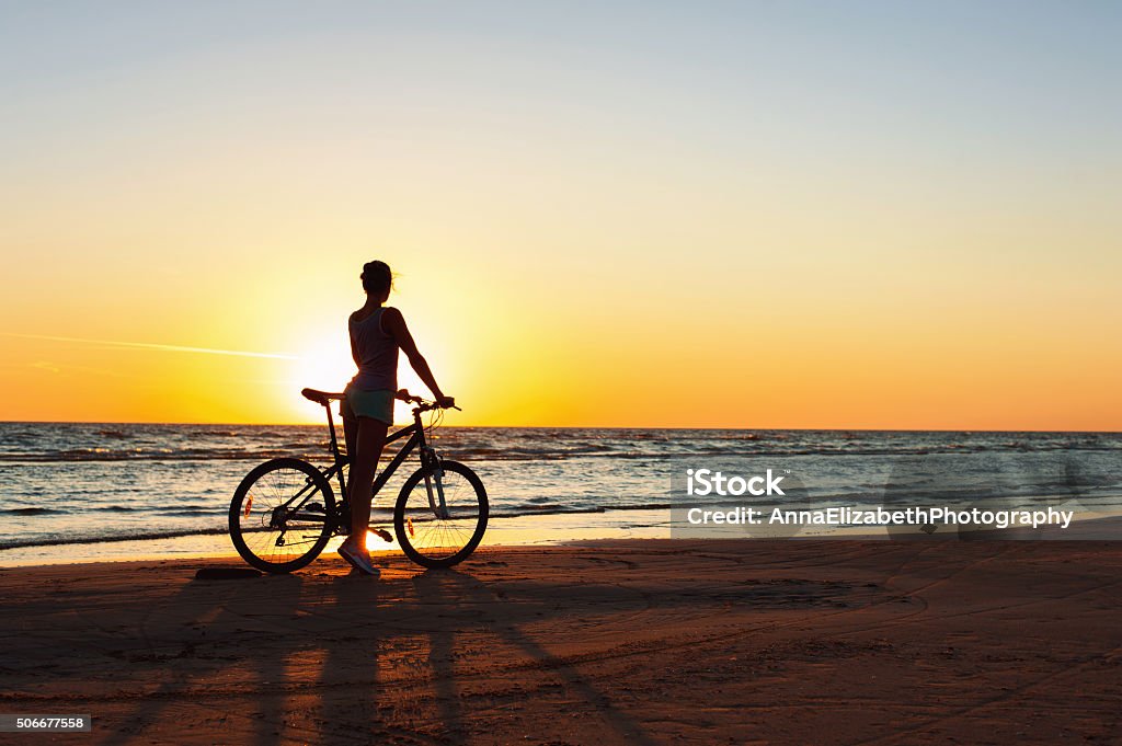 Catching a moment in time. Sporty woman cyclist at sunset Catching a moment in time. Young sporty woman cyclist silhouette contemplating the sunset on blue sky background on the beach. Summertime multicolored outdoors horizontal image with filter. Cycling Stock Photo