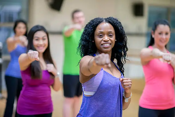 A multi-ethnic group of adults are taking a kickboxing class at the gym. One woman is smiling and looking at the camera.