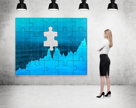 businesswoman standing with arms crossed in front of a puzzle on the concrete wall with a picture of blue graphs, one part missing, four lamps on the ceiling. Concept of getting the full picture.