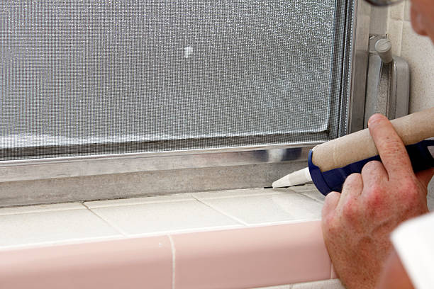 Caulking a Bathroom Window Frame Caucasian male hand holding a blue caulking gun about to caulk a crack between an interior gray aluminum metal window frame and a speckled off white tile base of an old bathroom window sill in the day. winterizing stock pictures, royalty-free photos & images