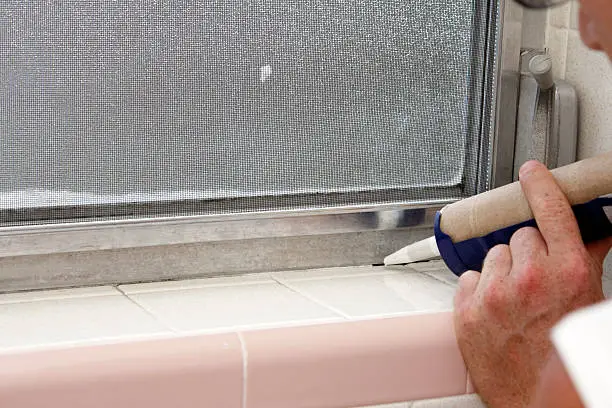 Caucasian male hand holding a blue caulking gun about to caulk a crack between an interior gray aluminum metal window frame and a speckled off white tile base of an old bathroom window sill in the day.