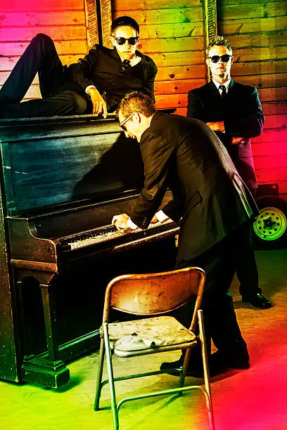 Three men in black suite wearing sunglasses at piano.  One playing, singing.  Others with serious looks, security staff. Colorful lights. Nightclub scene.   