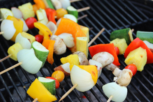 Seven (7) skewers of vegetables (includes bell pepper, mushroom, onion, zucchini, cherry tomato, pineapple, squash, and potato) on grill grate. Gray and glowing coals are visible beneath grate.