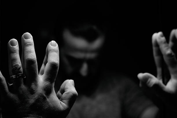 Man in despair with raised hands and bowed head Man in despair with raised hands and bowed head, monochromatic image in a low light room looking in front of mirror gloomy mood stock pictures, royalty-free photos & images