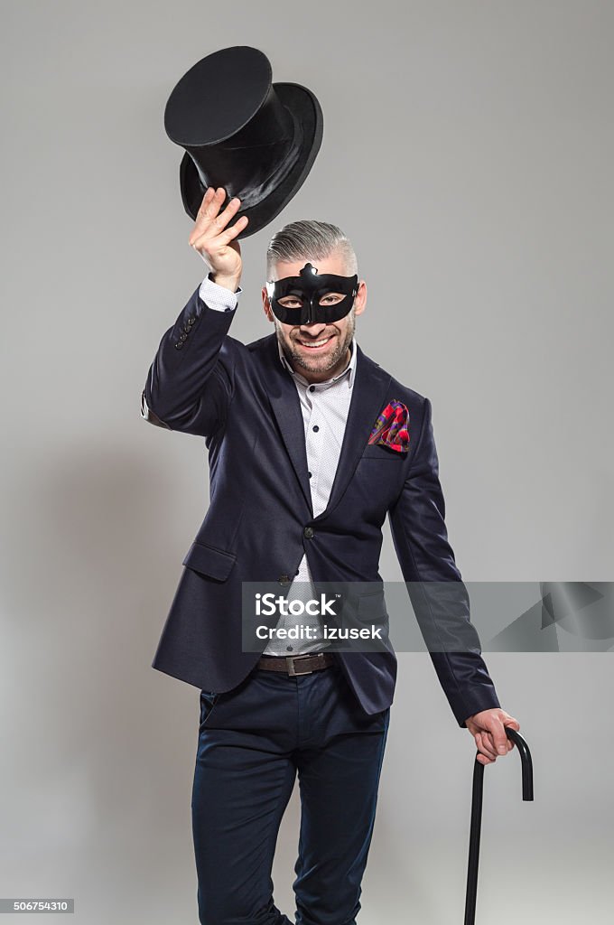 Wizard, elegant bearded man wearing top hat and carnival mask Portrait of elegant bearded businessman wearing jacket, top hat and carnival mask. Standing against grey background, holding walking stick in hand, raising his hat and smiling at camera. Studio shot, one person.  Top Hat Stock Photo