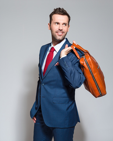 Portrait of elegant businessman wearing elegant suit, holding a leather briefcase, looking away. Studio shot, one person, grey background.