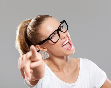 Portrait of excited young blond hair woman wearing white t-shirt and nerd glasses, pointing at camera. Focus on face. Studio shot, gray background.
