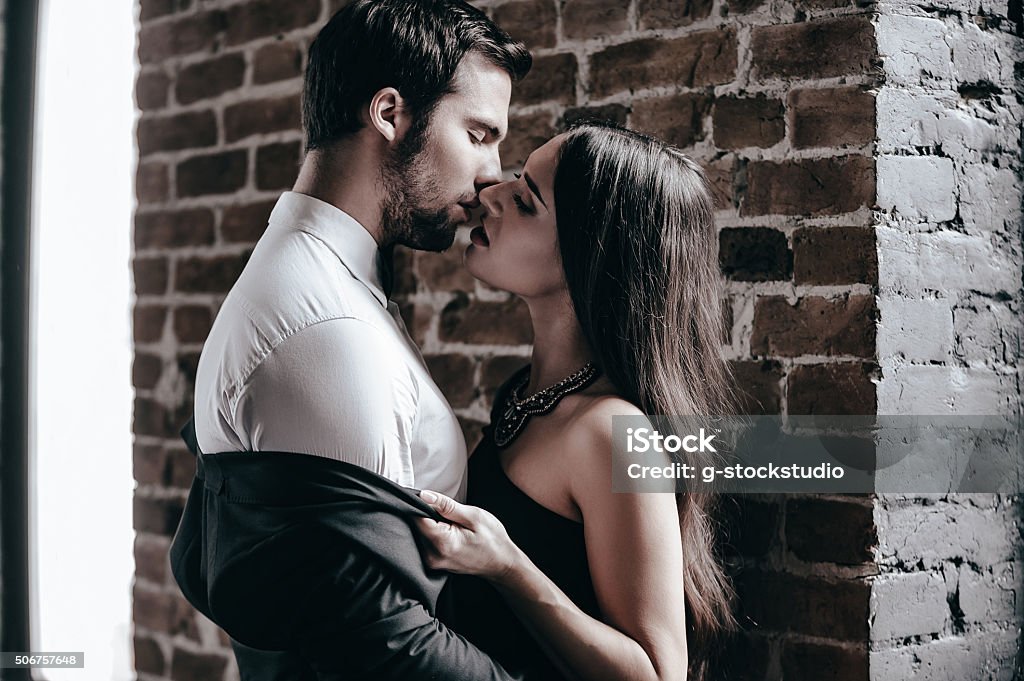 Stealing a kiss. Close-up side view of beautiful young woman in cocktail dress kissing her boyfriend and taking off his jacket while standing near brick wall in loft interior Couple - Relationship Stock Photo