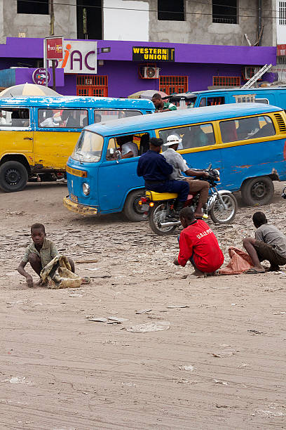 Children Collecting Rocks Kinshasa, Democratic Republic of the Congo - June 09, 2015: poor Children collecting stones from the ground whilst a motorcycle taxi and public buses go by. In the background, there's an unfinished building decorated purple on the ground floor with a Western Union sign above the door.  kinshasa stock pictures, royalty-free photos & images