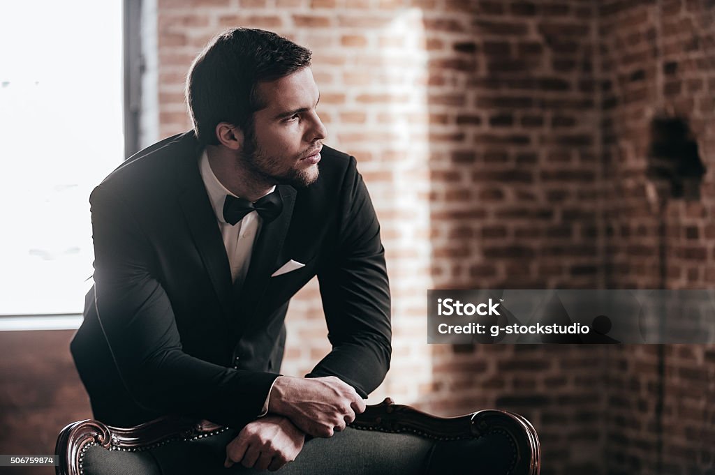Mr. Perfect. Thoughtful young handsome man in full suit and bow tie leaning to the chair and looking away while standing in loft interior Evening Wear Stock Photo