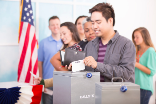 Multi-ethnic group of people stand in line to cast their ballot in the November USA elections at a local polling station. Asian man foreground. Other voters in line background. Ballot box.