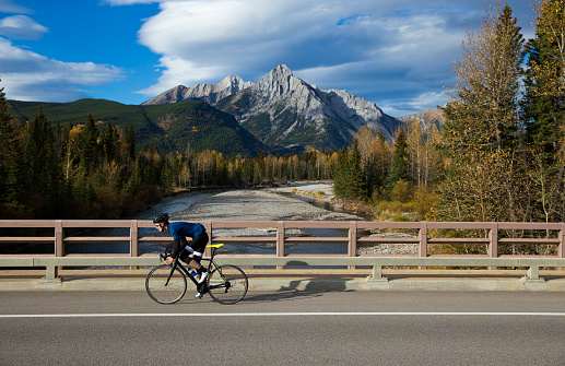 A male road cyclist rides on a bridge over a river in the Rocky Mountains of Canada.