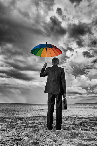 rear view of business man wearing suit, holding briefcase and protecting from the rain with colorful umbrella.economic crisis conceptual photo.
