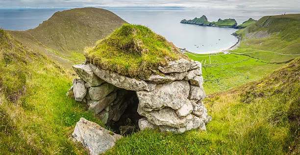 A traditional stone cleit, or store room, with turf roof to keep the rain off the peat, seabirds and grain stored inside overlooking the famous Village street and its cottages around the bay of Hirta, the remote island UNESCO World Heritage Site in the St. Kilda archipelago with the mountain peaks of the Western Isles far away on the eastern horizon, Scotland. ProPhoto RGB profile for maximum color fidelity and gamut.
