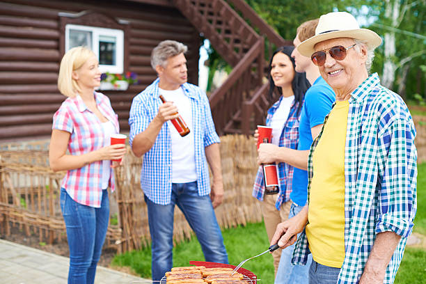 Portrait of happy senior man frying sausages and looking at in the countryside at weekend with group of young friends talking on background