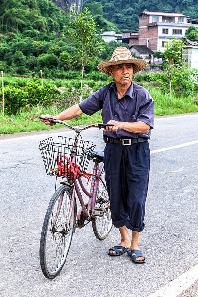 Yong Chung, China - May 5, 2015: Chinese farmer in a straw hat with bicycle stands on the road. In the background is visible Yong Chung village in Southern China, about 50 km from Guilin.