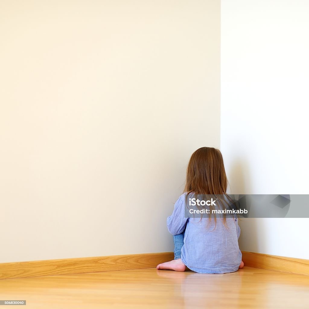 Sad little girl sitting in a corner Very sad little girl sitting on a floor at home Behind Stock Photo