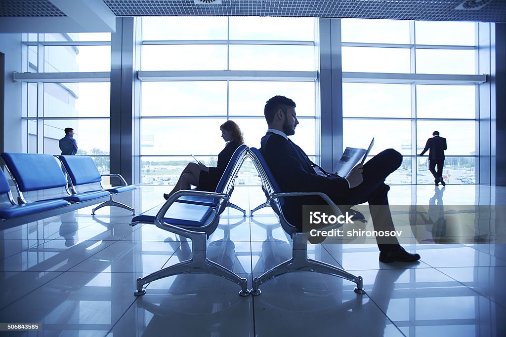 At the airport Two business partners reading at the airport on background of their colleagues by the window Adult Stock Photo