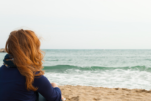 Redhead girl on a Barcelona sand beach looking at the sea, view from behind