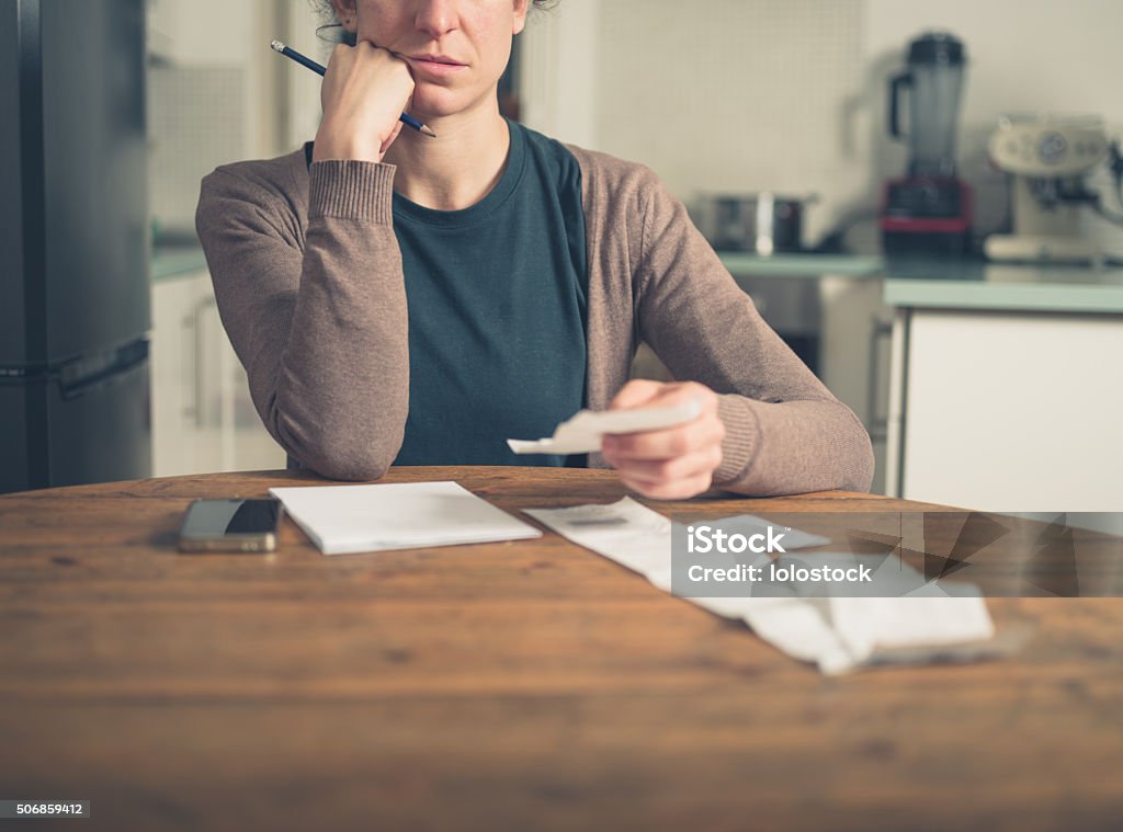 Woman looking at receipts at home A young woman is sitting in her kitchen and is loking at her receipts at home while using a smart phone Currency Stock Photo