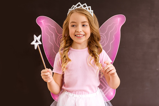 Young queen fairy with pink wings holding a magic wand against dark background