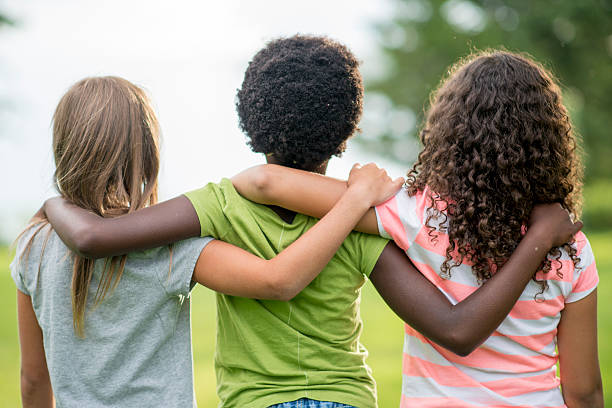 Friends Standing Together A rear view of a multi-ethnic group of elementary age girl friends are standing together with their arms around each other. They are looking out at the park on a spring day. only girls stock pictures, royalty-free photos & images