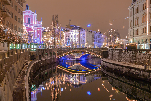 Ljubljana, Slovenia - January 3, 2016. Snowy banks of Ljubljanica river, Triple bridge, on the background with illuminated Franciscan Church of the Annunciation and christmas tree.