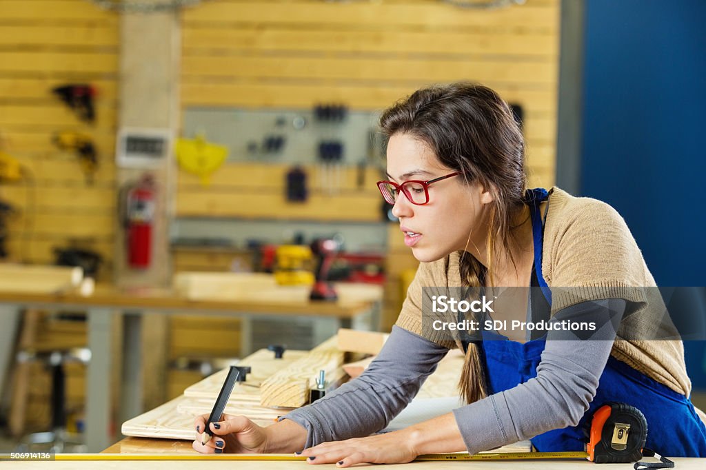 Female carpentry apprentice measuring boards in professional workshop or makerspace Young adult Caucasian woman is apprentice in professioal carpentry workshop or makerspace. Worker is measuring boards to cut while building furniture or cabinetry for a customer. Accuracy Stock Photo