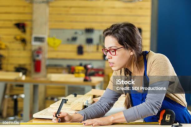 Female Carpentry Apprentice Measuring Boards In Professional Workshop Or Makerspace Stock Photo - Download Image Now