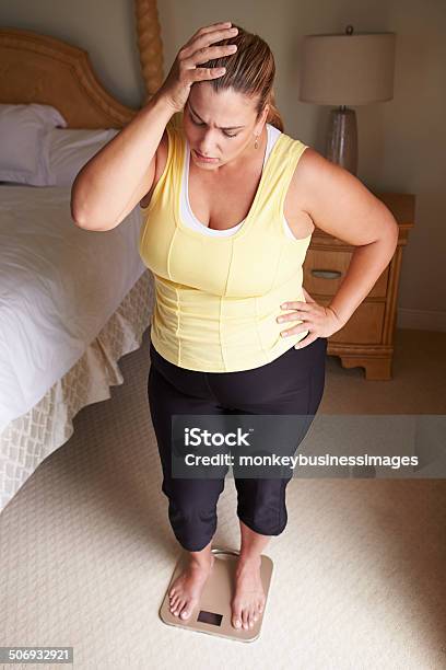 Overweight Woman Weighing Herself On Scales In Bedroom Stock Photo - Download Image Now