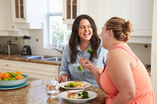Two Overweight Women On Diet Eating Healthy Meal In Kitchen Sitting Down Laughing