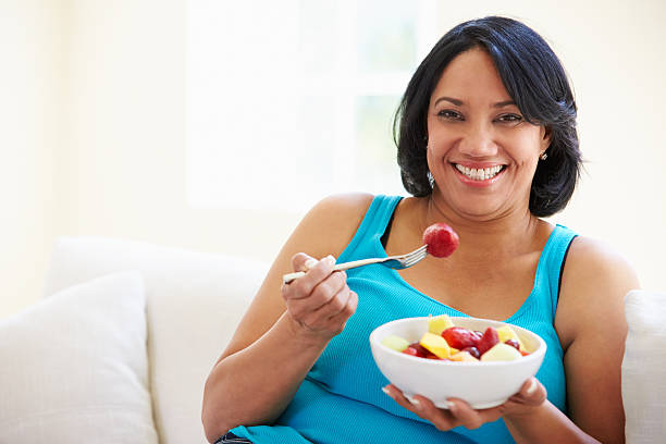 Overweight Woman Sitting On Sofa Eating Bowl Of Fresh Fruit Overweight Woman Sitting On Sofa Eating Bowl Of Fresh Fruit At Home Looking Happy fat nutrient photos stock pictures, royalty-free photos & images