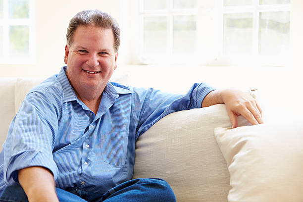 Portrait Of Overweight Man Sitting On Sofa Portrait Of Overweight Man Sitting On Sofa Relaxing Whilst Smiling At Camera overweight man stock pictures, royalty-free photos & images