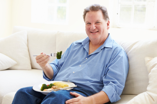 Overweight Man Eating Healthy Meal Sitting On Sofa Looking To Camera Smiling