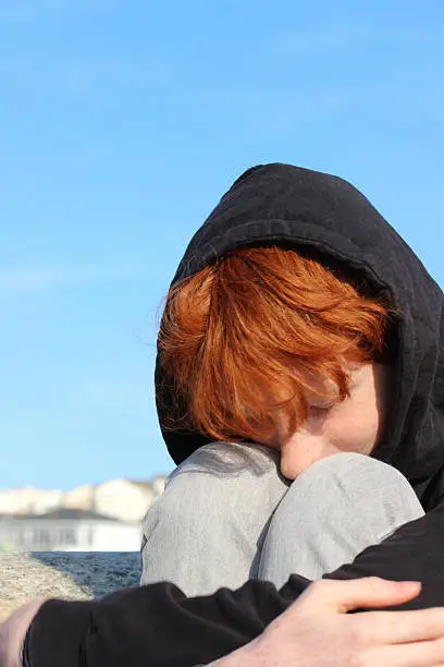 Photo showing a teenager daydreaming at the beach, looking down at the sand.  This teenage boy has red hair and is pictured sitting whilst being curled up, in a foetus position, with his head in his knees.  The sea and seaside resort form the blurred background on this sunny summer's day.