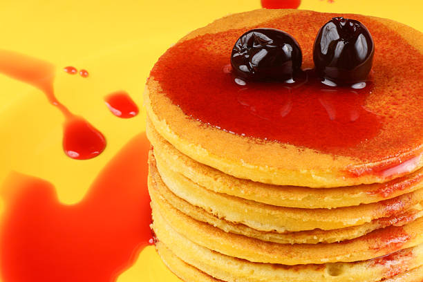 Close-up of a stack of corn pancakes with syrup and sour cherries on a yellow plate.