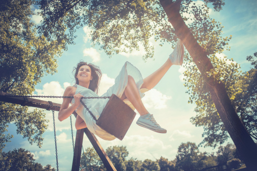 A happy young woman laughing on a bright sunny day while having fun on a swing in the park.  She grins widely and kicks her legs in the air as she reaches the apex of her swing. The image is taken from below as she swings, and there are leafy trees and a blue sky with clouds in the background. Developed from RAW; retouched with special care and attention; Small amount of grain added for best final impression; Ready made for print and web use;