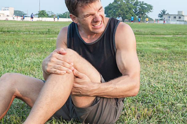 Leg injury Man suffers painful hamstring injury and holds painful area with hands as he sits on grassy ball field hamstring injury stock pictures, royalty-free photos & images