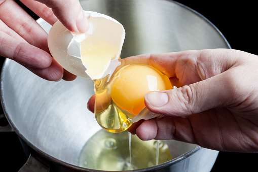 Separating yolk and white of the white shell egg in woman’s hands above metal stainless steel mixing bowl on black background