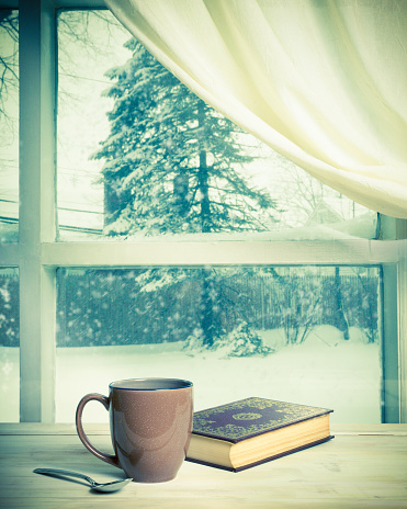 Cup of coffee and book near window on a snowy winter day