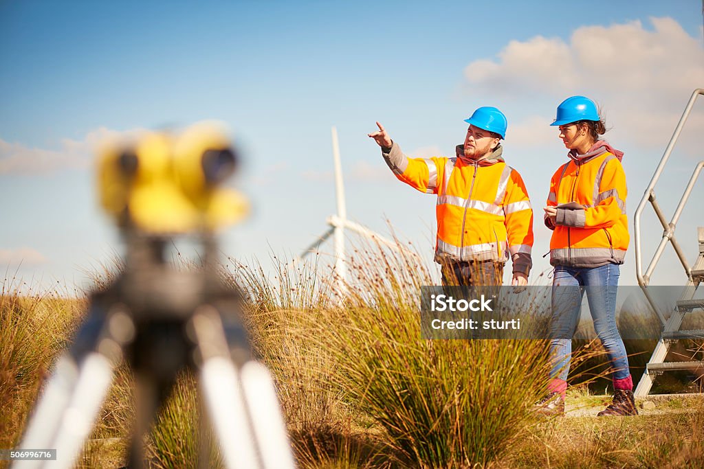 male and female windfarm engineers two wind farm engineers using a builder's level to plan out the expansion of the wind farm site. they are wearing orange hi vis jackets and blue hard hats . one is male , one is female. In the foreground the female is looking through the level whilst the male engineer is approaching .In the background wind turbines can be seen across the landscape. Construction Industry Stock Photo