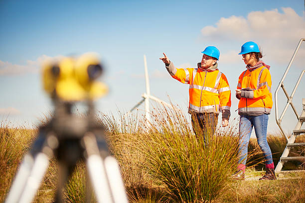male and female windfarm engineers two wind farm engineers using a builder's level to plan out the expansion of the wind farm site. they are wearing orange hi vis jackets and blue hard hats . one is male , one is female. In the foreground the female is looking through the level whilst the male engineer is approaching .In the background wind turbines can be seen across the landscape. environmentalist photos stock pictures, royalty-free photos & images