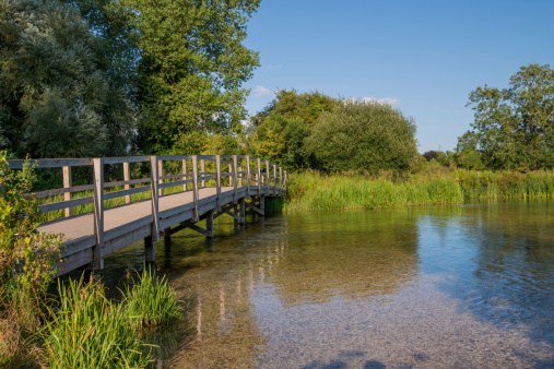 A wooden footbridge crosses the River Test in Houghton, Hampshire, England. It is a sunny summer afternoon with the bridge reflected in the river water.