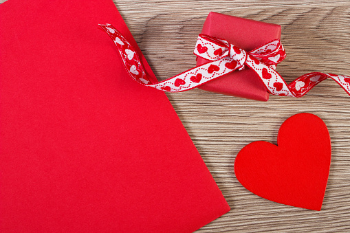 Wrapped gift with ribbon, red wooden heart and love letter in envelope on wooden background, decoration for Valentines Day, copy space for text