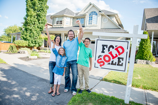 A family of four is happily standing outside of their new home next to a sold sign. They are smiling and looking at the camera.